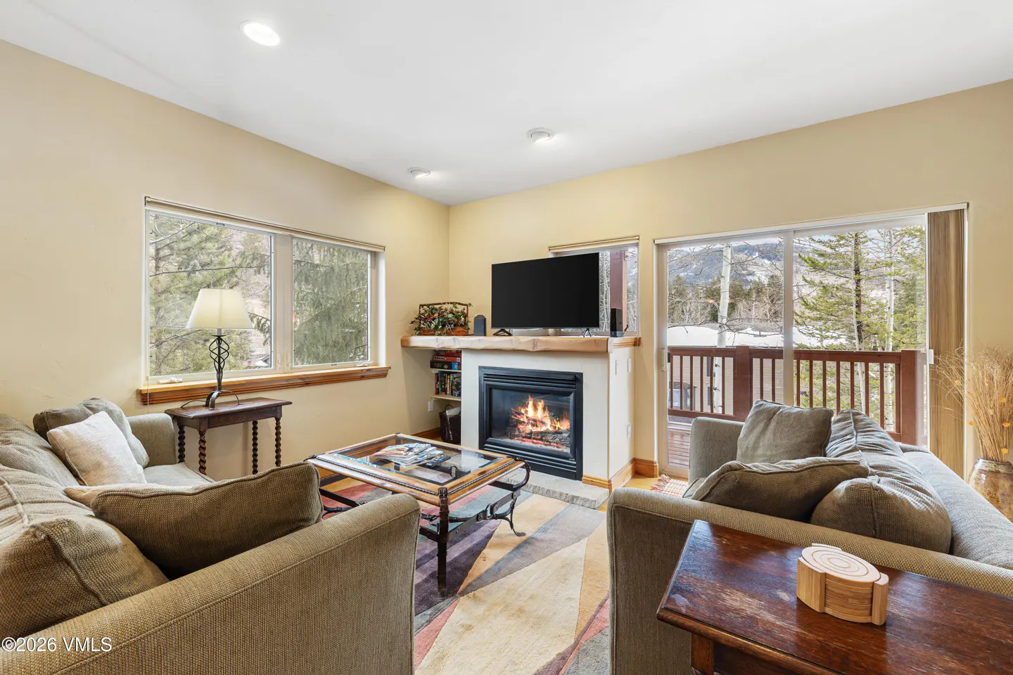 Living room with beige walls, two sofas, a fireplace with a TV above, and windows with views of trees and mountains.