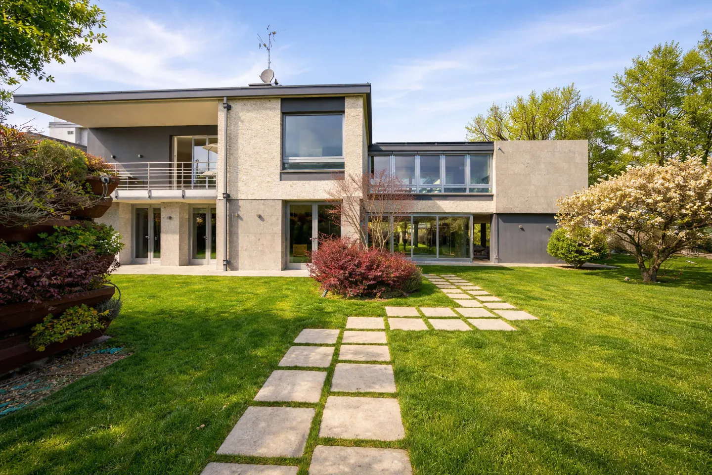 Modern two-story house with a stone walkway leading through a green lawn. Balcony, large windows, and trees visible.