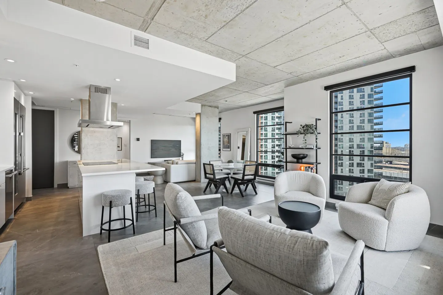 Open-concept condo with concrete ceilings, large windows, and city views. The living area features modern furniture and a white kitchen island.