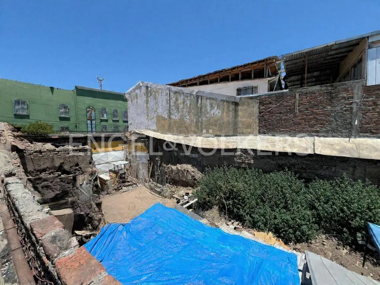 View of a vacant lot with exposed brick, covered by a blue tarp, surrounded by buildings under a clear blue sky.