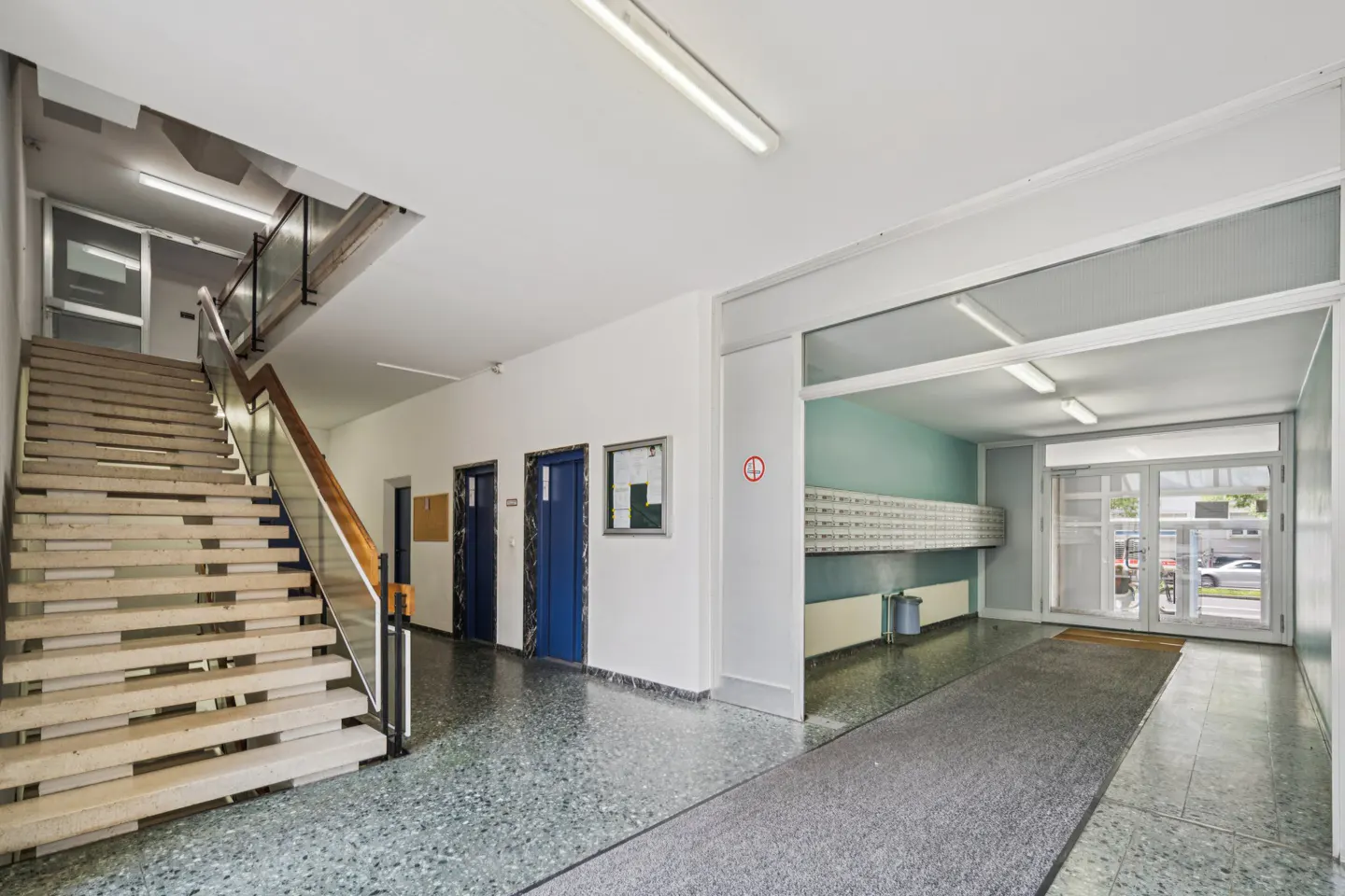Bright apartment building lobby with a staircase, two blue elevator doors, a row of mailboxes, and glass entrance doors.