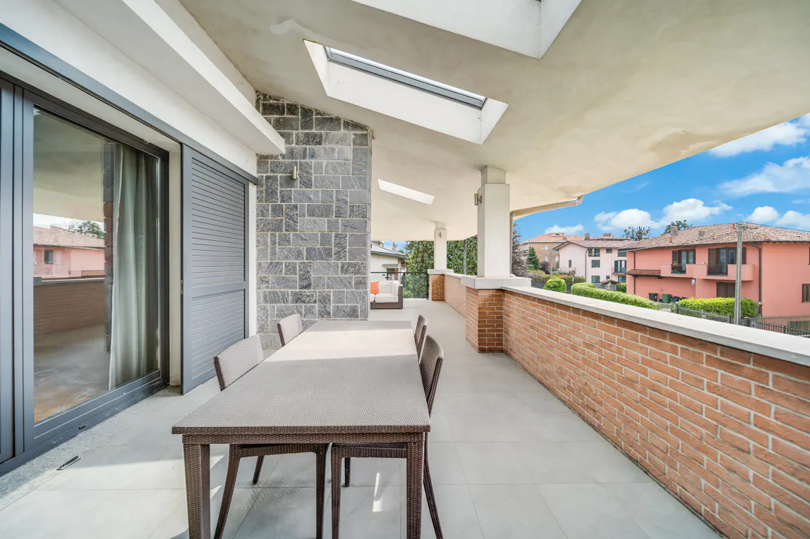 A covered patio with a table and chairs overlooks a neighborhood. The patio has a brick railing and a stone accent wall. Skylights are visible in the ceiling.