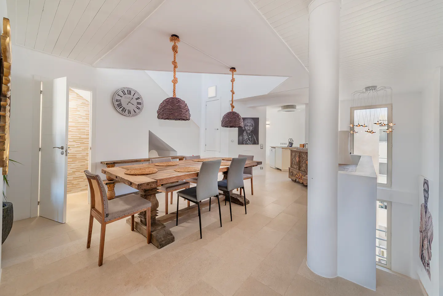 Bright, open dining area with a rustic wood table, gray chairs, and woven pendant lights. White walls, tile floors, and a large clock add to the airy feel.