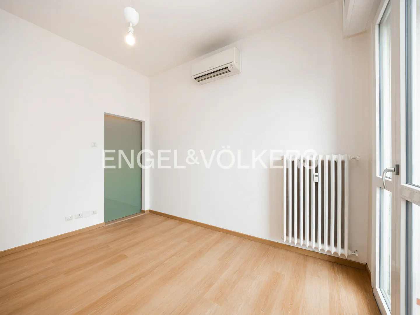 Bright, empty room with light wood floors, white walls, radiator, and glass door. Air conditioner on the wall.