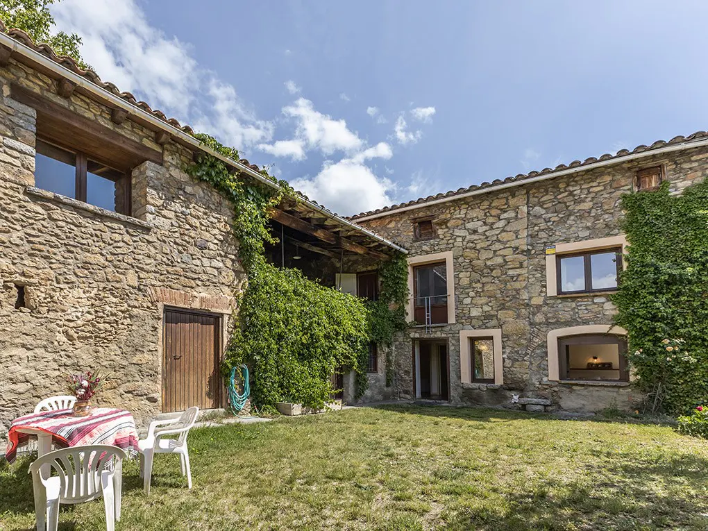 Stone house with green lawn, table, and chairs. Vines climb the walls under a blue sky with clouds.