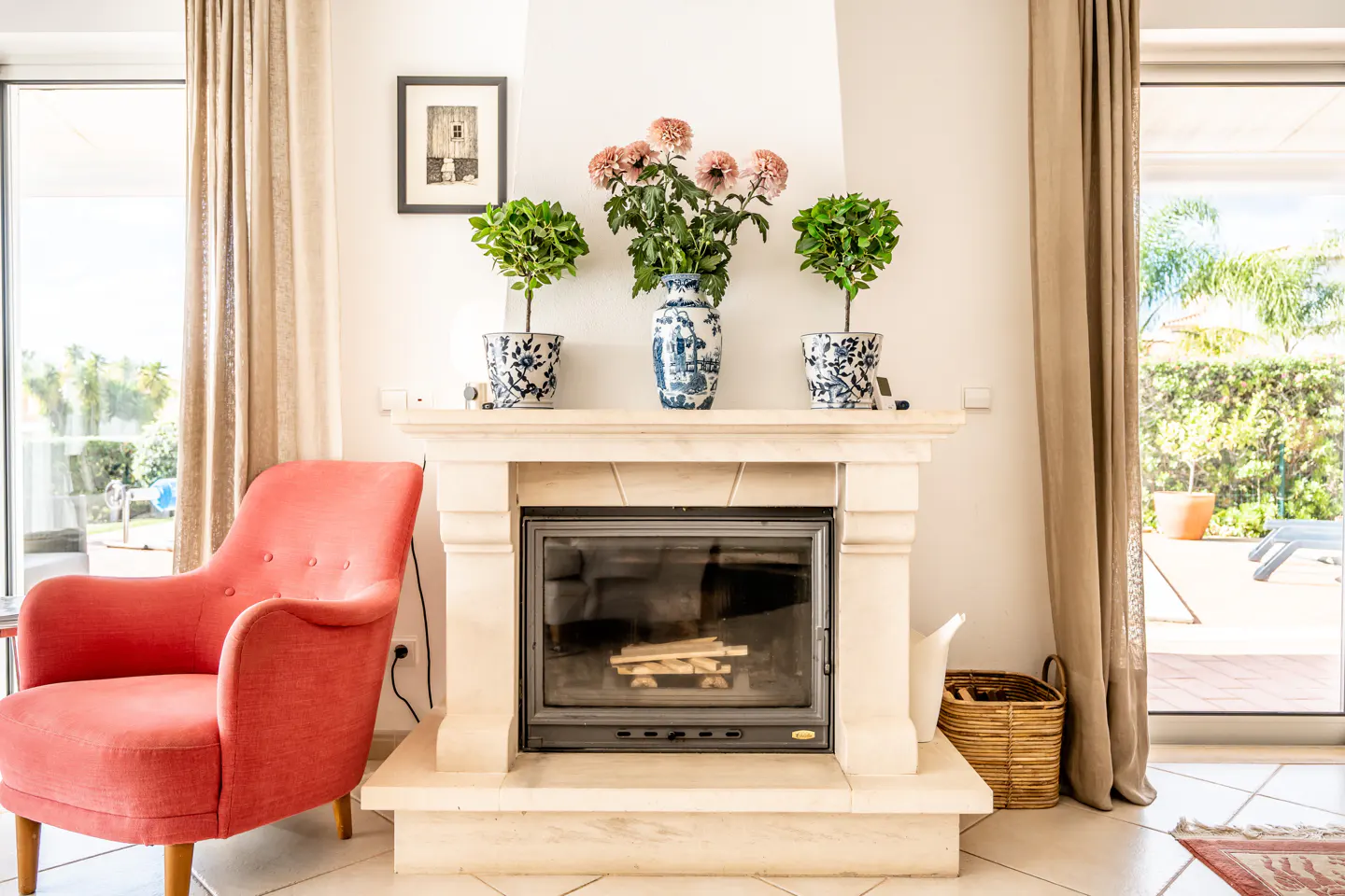 Living room with a stone fireplace, topped with plants and flowers. A coral armchair sits to the left, and large windows offer a view of the outdoors.