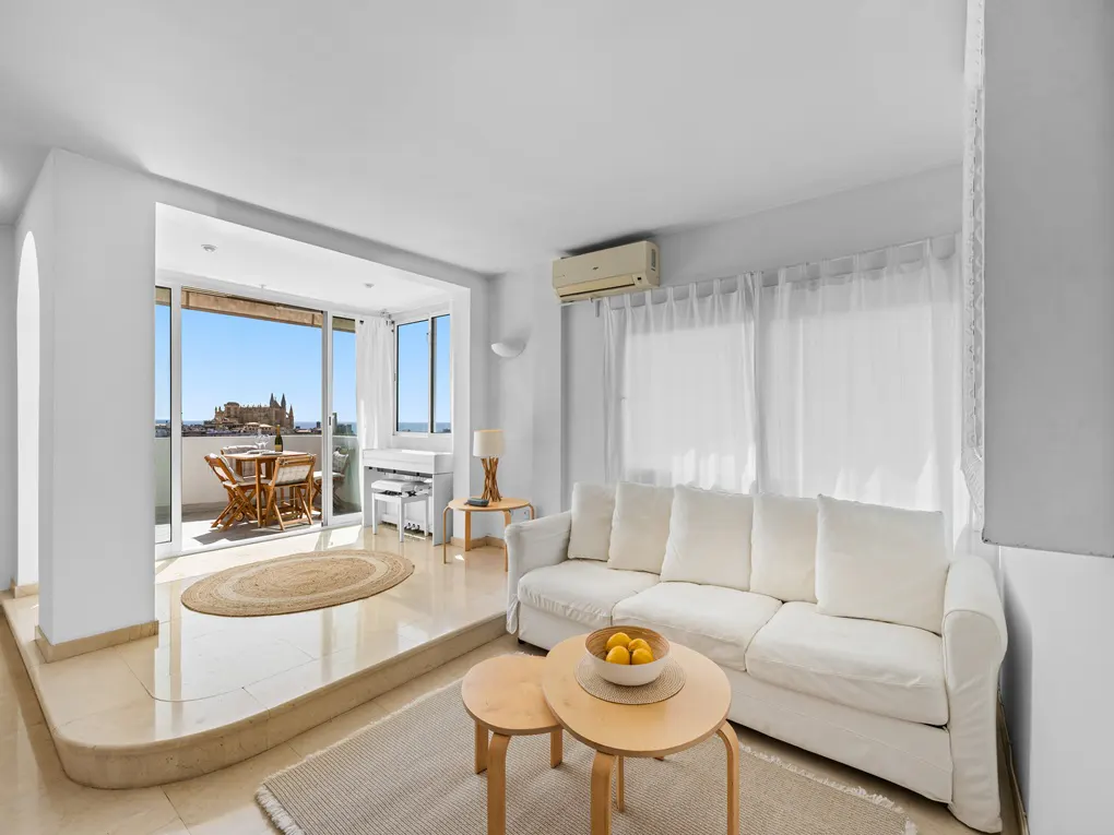 Bright living room with white sofa, round tables, and balcony. Outdoor seating and a distant cathedral are visible.