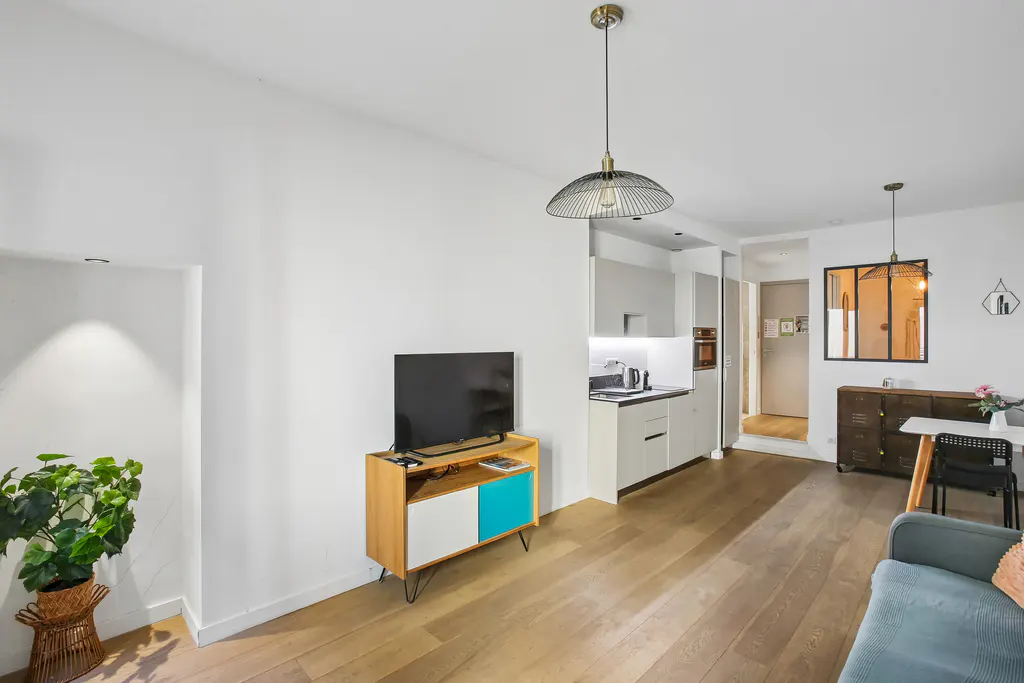 Bright apartment interior with wood floors, white walls, and modern furnishings. A TV sits on a cabinet with blue accents, near a compact kitchen.