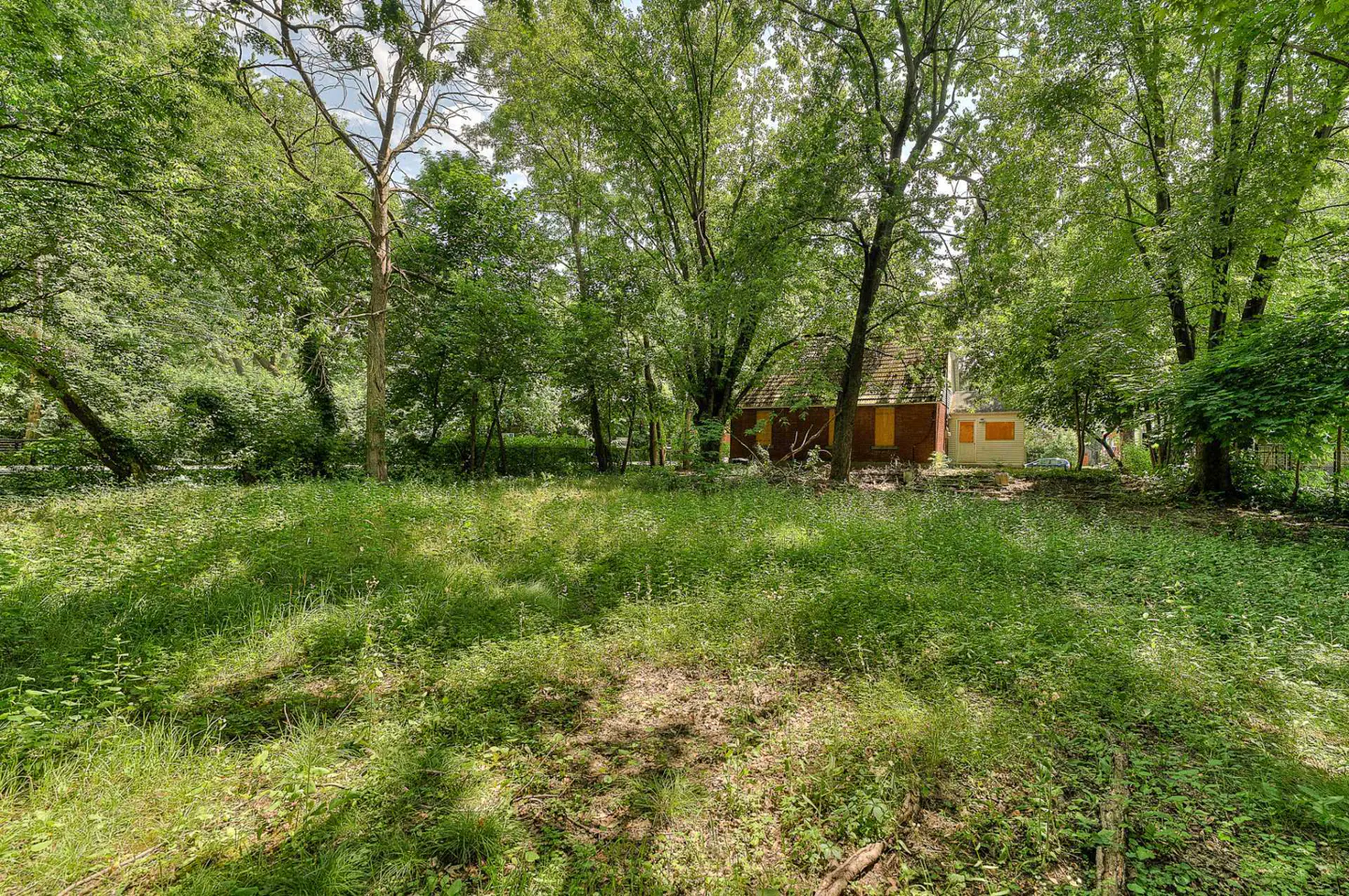 Overgrown lawn with trees leading to a red brick house with boarded windows in the background.