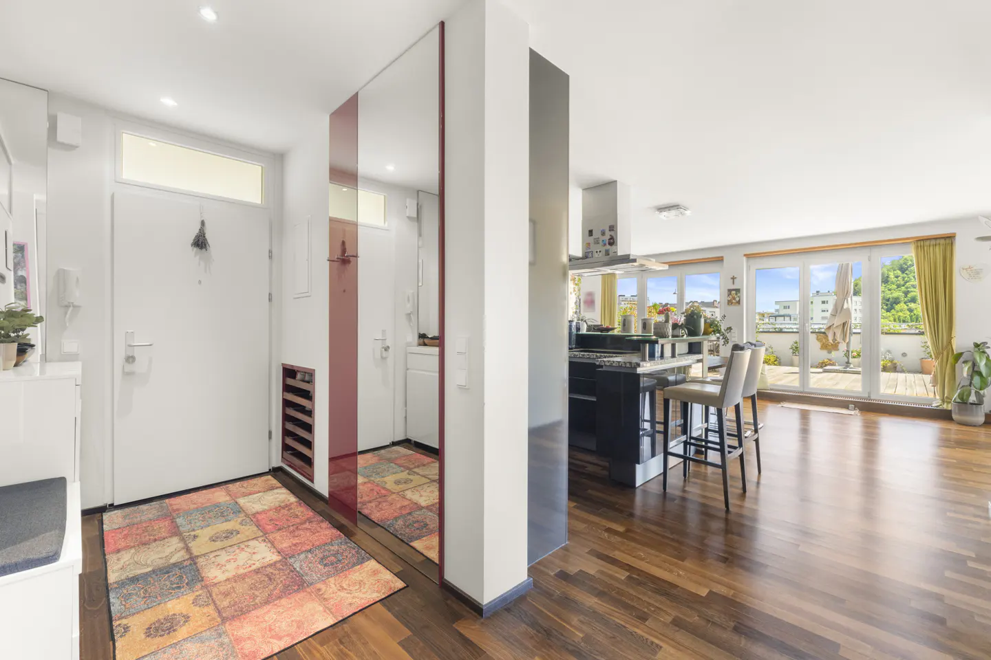 Bright apartment interior with hardwood floors, a colorful rug, and a kitchen island with bar stools. Balcony doors open to a view.