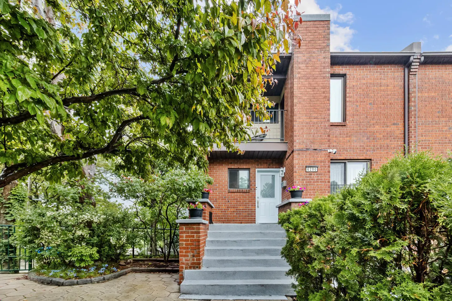 A red brick townhouse with a white front door, gray concrete steps, and lush green trees and shrubs in the front yard.