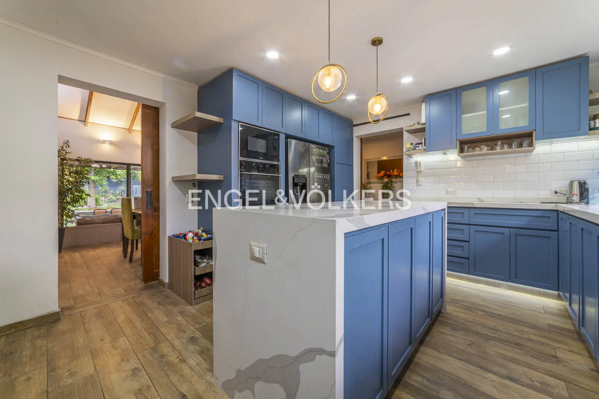 A modern kitchen with blue cabinets, a white marble island, and wood floors. Two pendant lights hang above the island. A doorway leads to a living room.