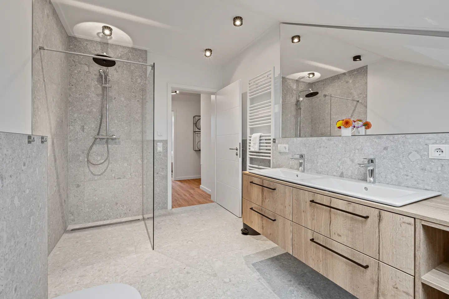 Modern bathroom with gray stone tile, glass shower, and wood vanity with double sinks. A white door is slightly ajar.