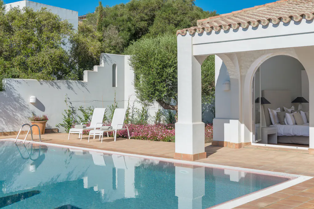 A turquoise pool reflects a white villa with arched openings and a glimpse of a bedroom. Two white lounge chairs sit nearby.
