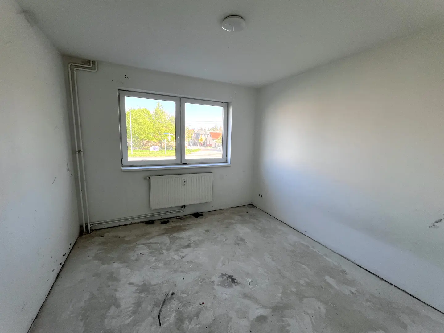 Empty room with white walls, a window showing trees, and a concrete floor. A radiator is under the window.