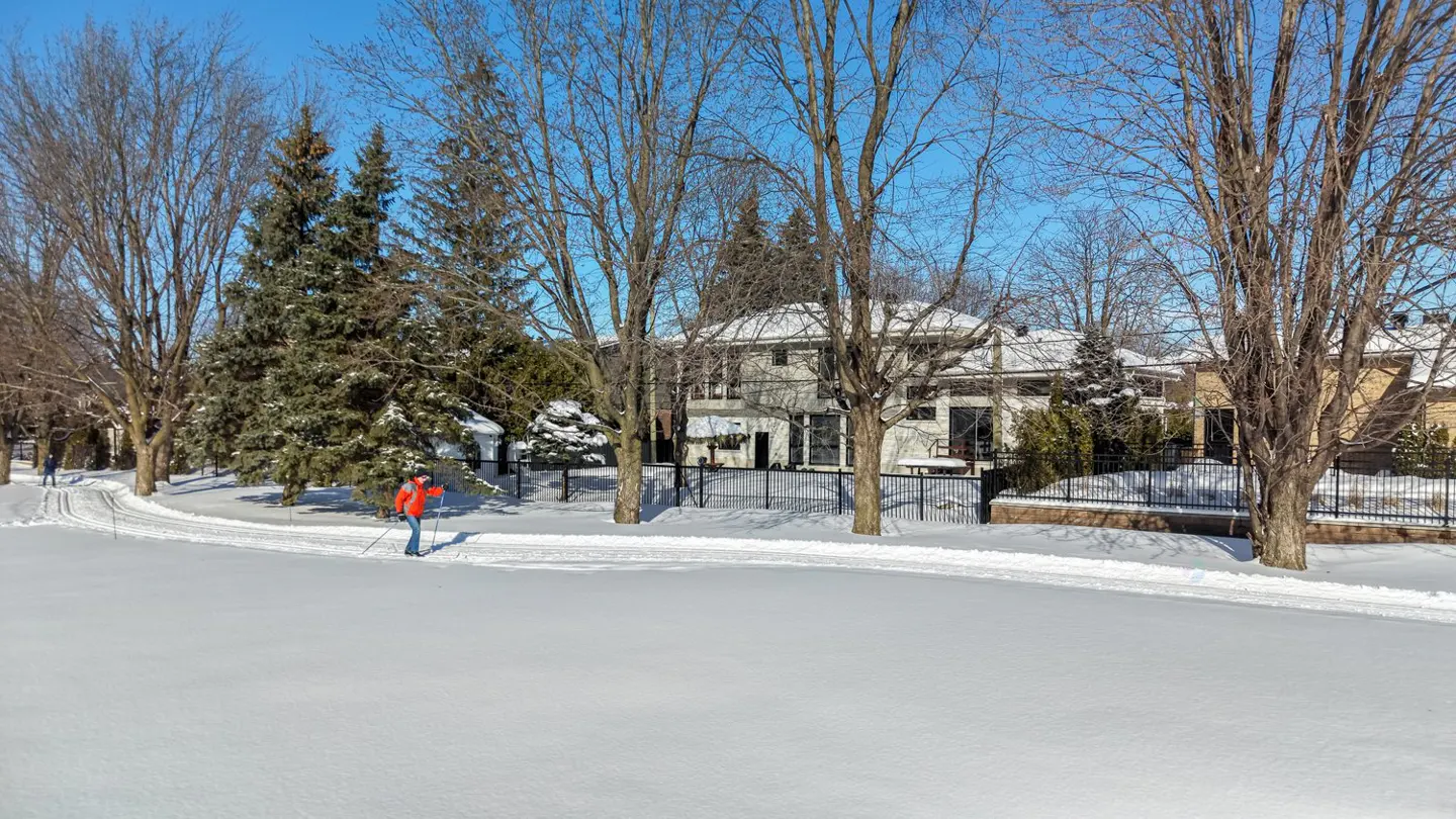 A person cross-country skis on a snowy path in front of a house on a sunny winter day.