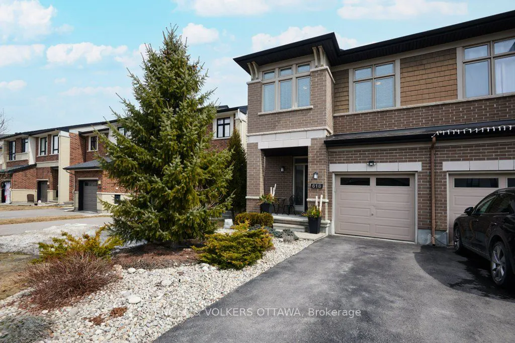 Exterior view of a two-story brick townhouse with a beige garage door and a black car parked in the driveway. A tall evergreen tree stands in the front yard.