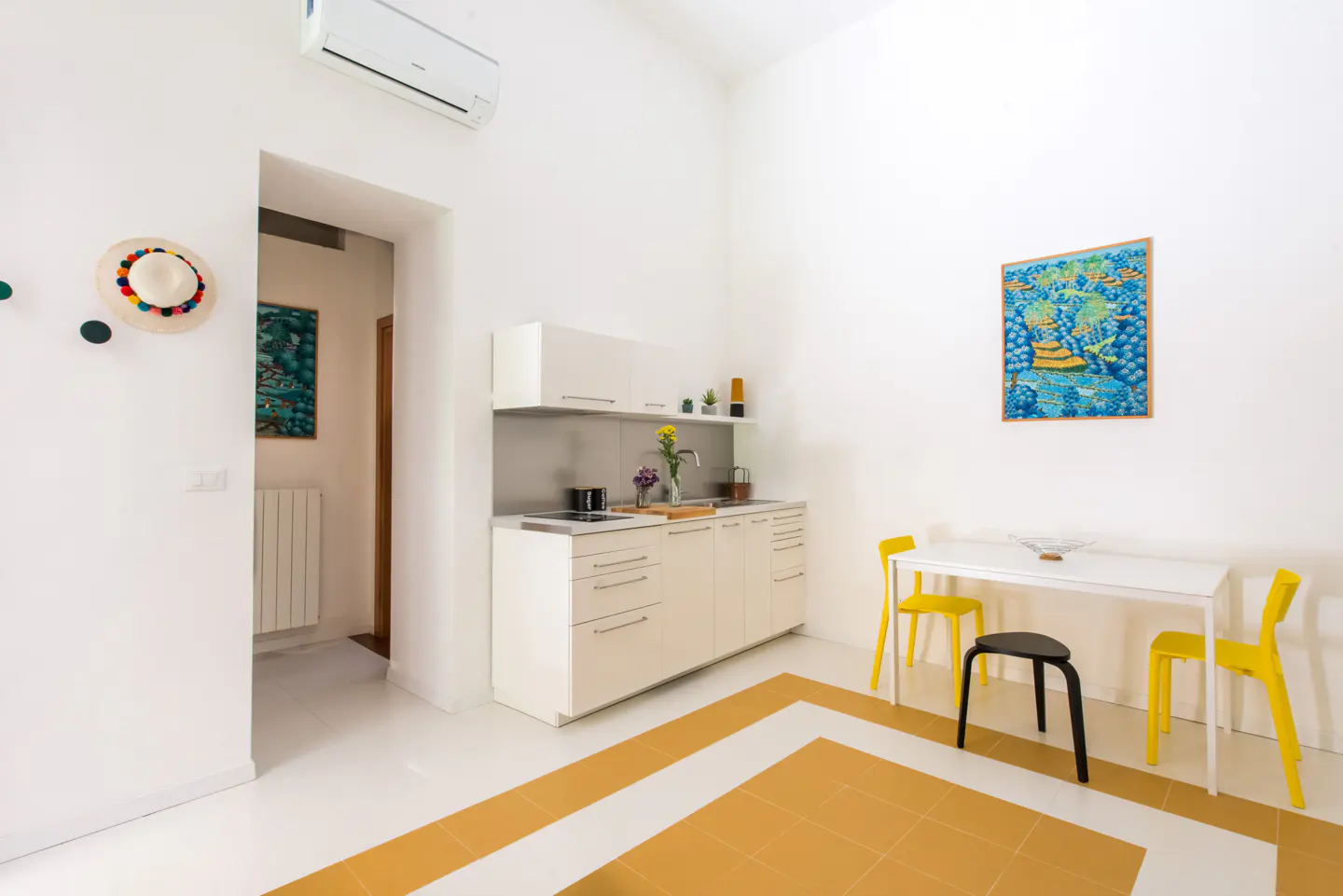 Bright, white apartment interior with a small kitchen, dining table with yellow chairs, and yellow-and-white tiled floor. A colorful hat hangs on the wall.