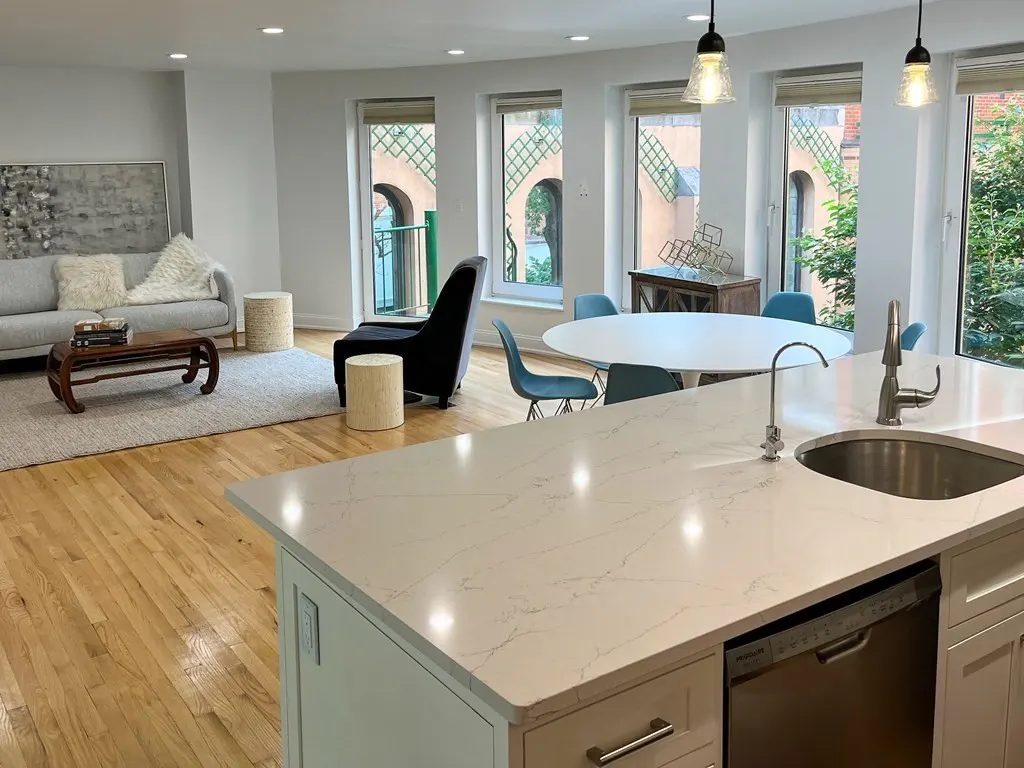 Open-concept living space with a kitchen island, hardwood floors, and large windows. A gray sofa and round table are visible.