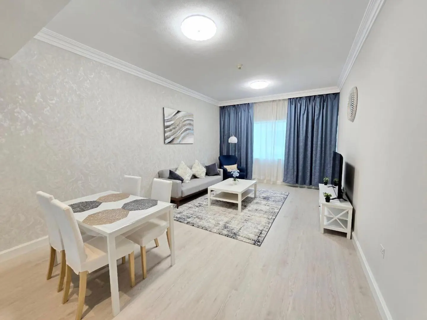 Bright, modern living room with a white dining table, gray sofa, blue curtains, and a patterned rug. A TV sits on a white stand.