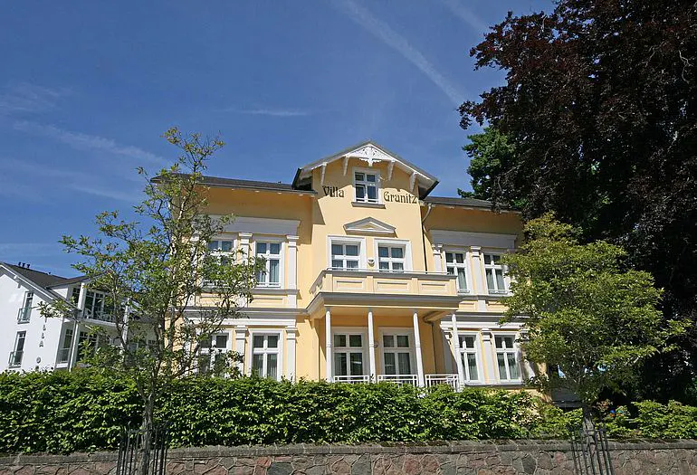 A pale yellow historic villa named Villa Granitz with white trim, a balcony, and a stone wall under a clear blue sky.