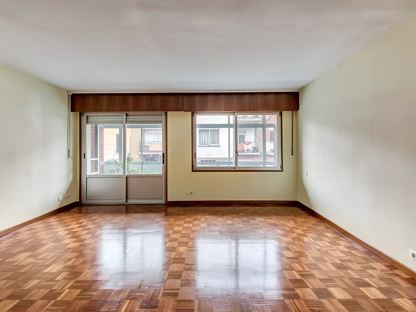 Empty room with parquet floor, light yellow walls, and two windows with brown trim. Outside view of buildings and greenery.