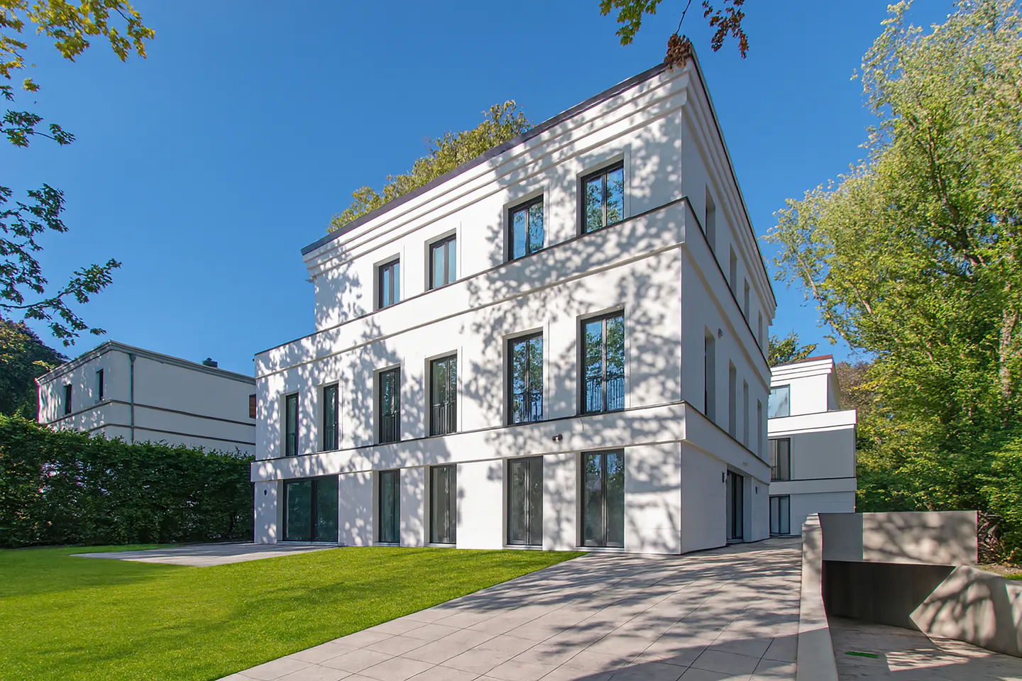 A modern, three-story white building with black-framed windows, a green lawn, and a paved driveway under a clear blue sky.