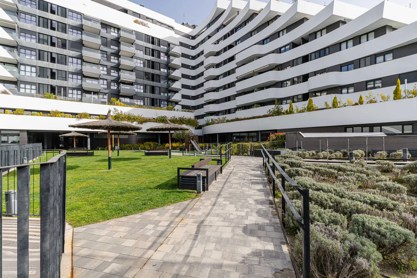Exterior view of a modern apartment building with a courtyard, featuring grass, benches, and straw umbrellas.