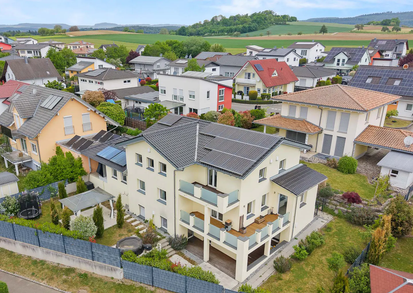 Aerial view of a two-story, cream-colored house with a gray roof and solar panels in a suburban neighborhood.
