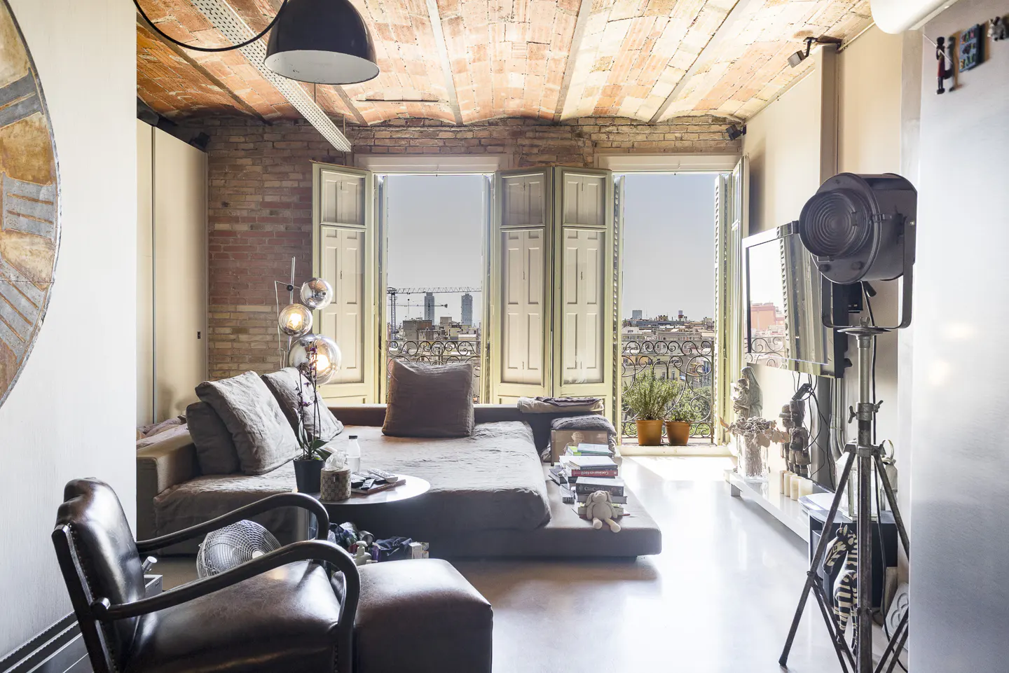 Living room with exposed brick walls and ceiling, a large sofa, and open shutters overlooking a city. A black spotlight stands near a TV.