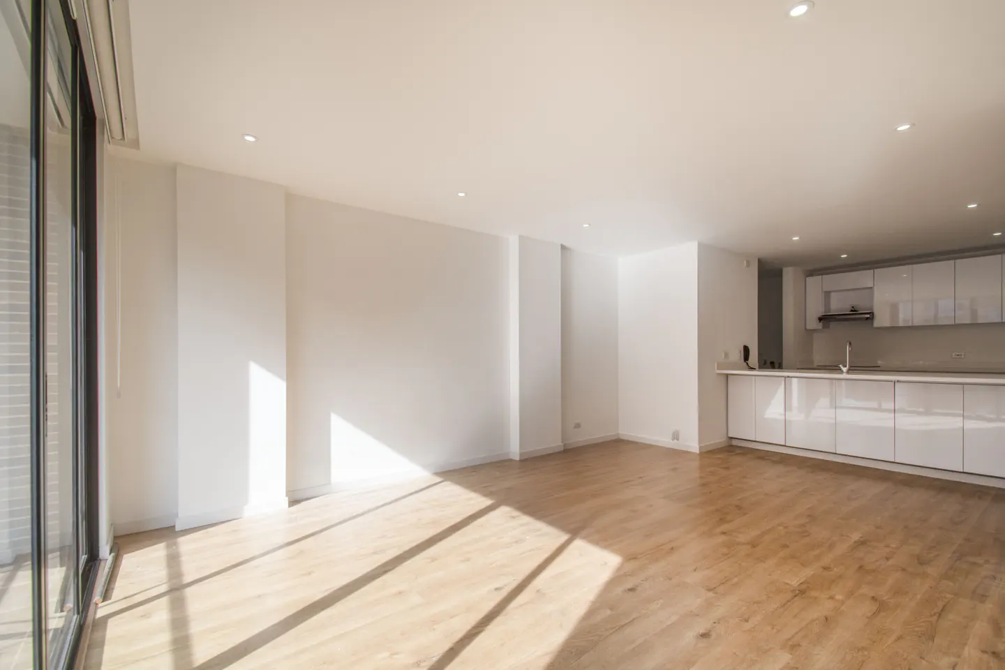 Bright, empty apartment with wood floors, white walls, and a modern white kitchen. Sunlight streams in from a large window.