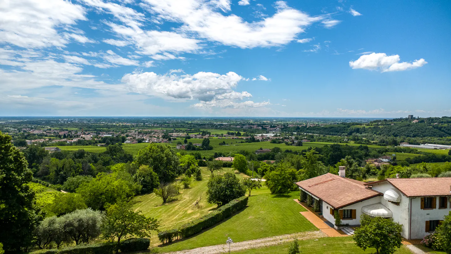 A white house with a terracotta roof sits on a lush green hillside overlooking a vast valley under a bright blue sky.