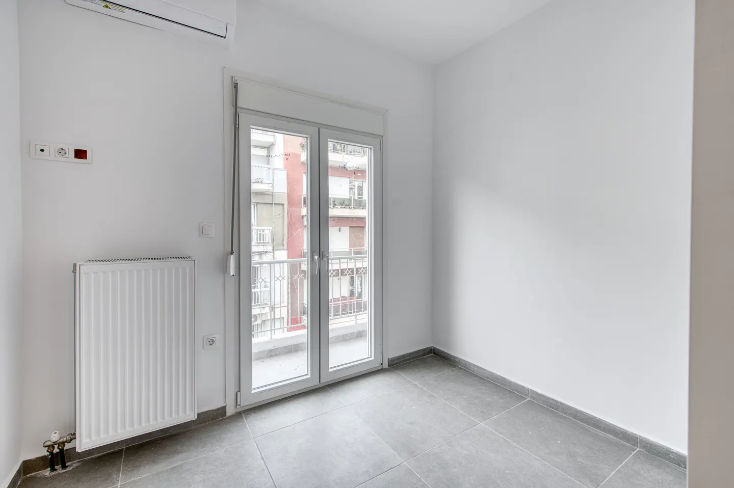 A bright, empty room with white walls, gray tile floor, and a white-framed glass door to a balcony. A white radiator is on the left wall.
