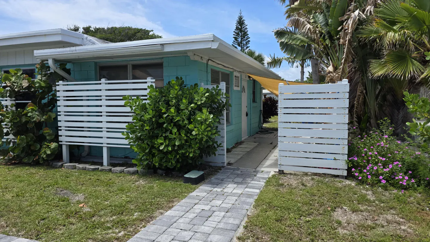 Exterior of a light blue beach house with a white fence, brick walkway, and tropical plants.