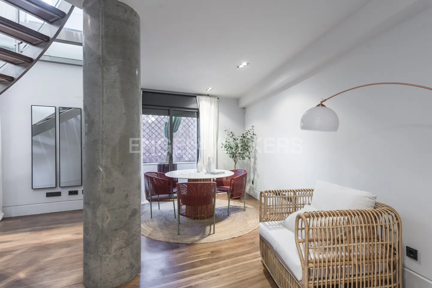 Bright living space with wood floors, a concrete pillar, and a round dining table with red chairs. A wicker chair sits near a white wall with a curved lamp.