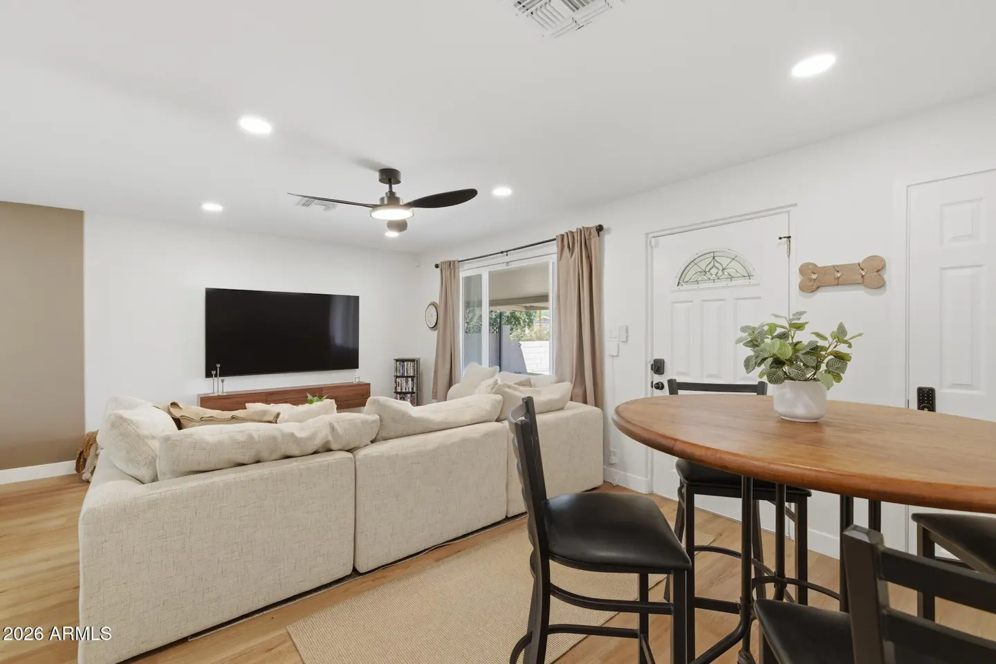 Bright living room with a large beige sectional sofa, wood floors, and a round wooden table with black chairs. A large TV is mounted on the wall.