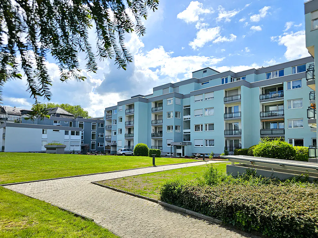 Exterior view of a light blue apartment building with balconies, green lawn, and a stone walkway.