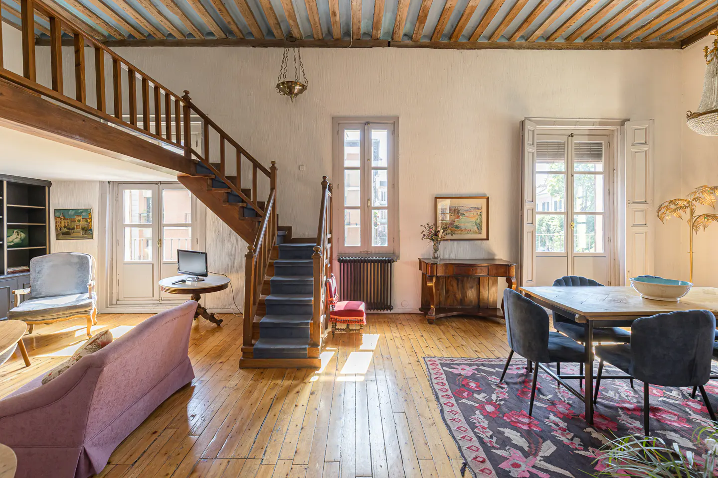 A bright living room with wood floors, a staircase, and a dining table with blue chairs.