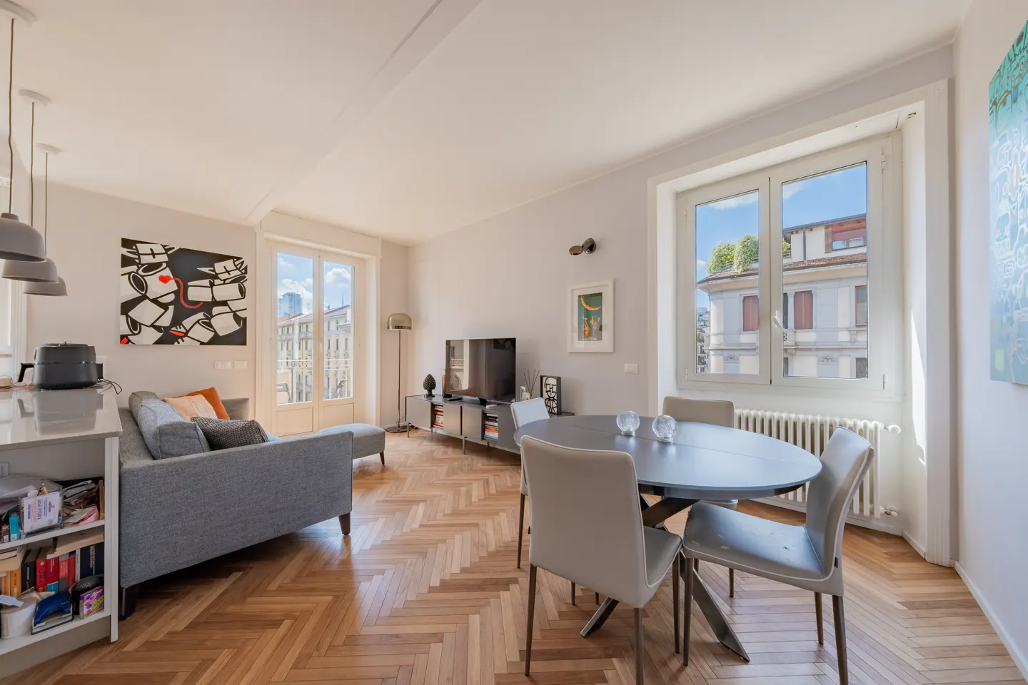 Bright living room with herringbone wood floors, gray sofa, round table with chairs, and city views through large windows.