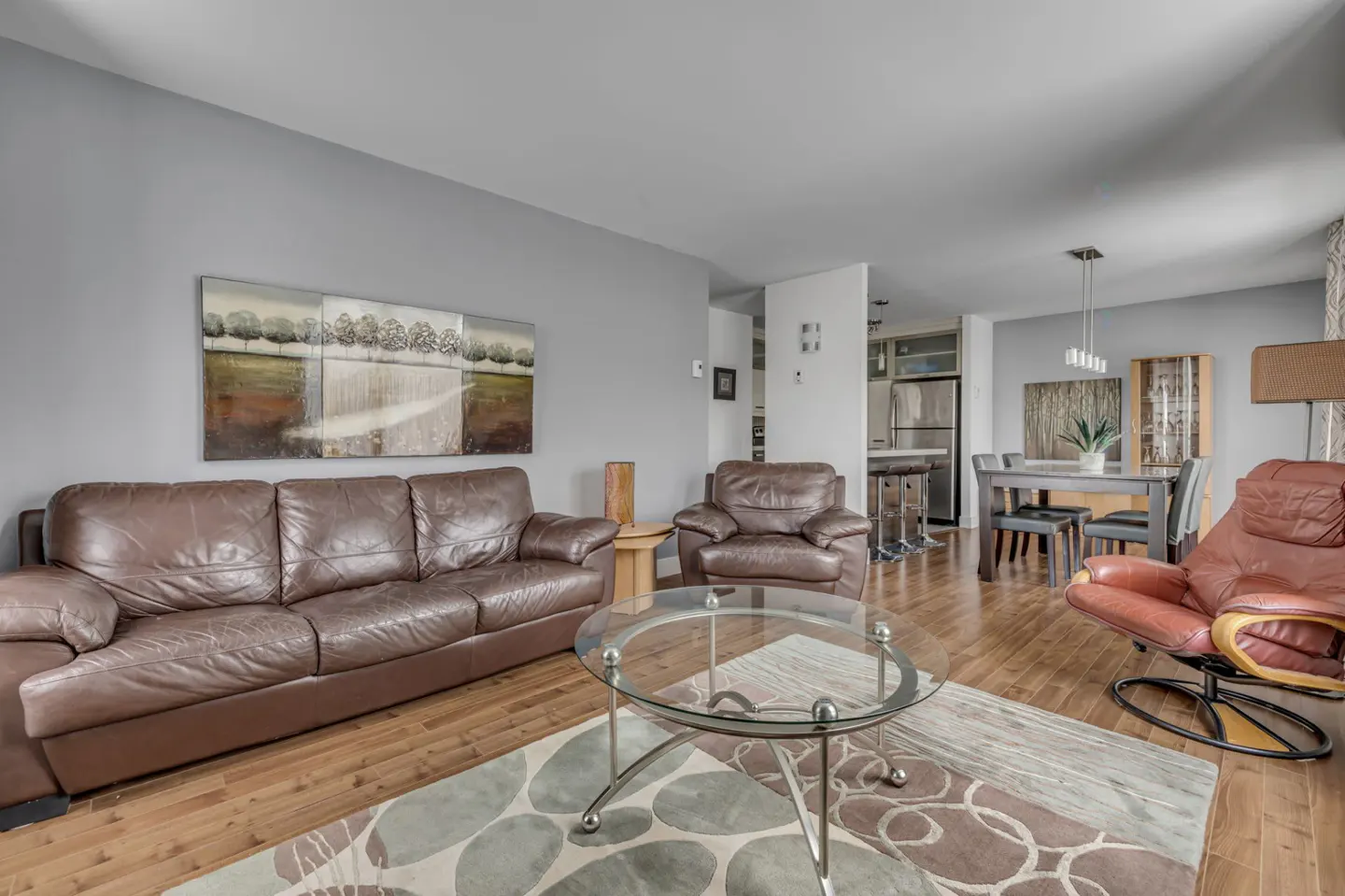 Living room with brown leather sofa, chair, and recliner. A glass table sits on a patterned rug. Dining area and kitchen are in the background.