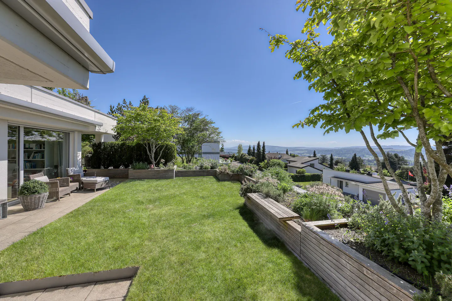 A modern home's backyard features a green lawn, wooden planters, and a patio with wicker furniture under a clear blue sky.