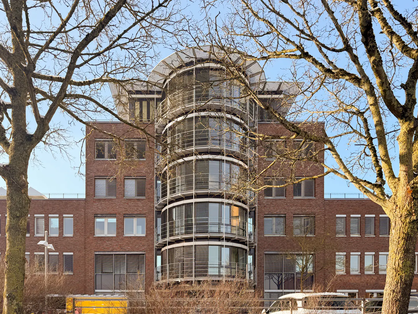 Brick building with a curved glass and metal facade, framed by bare trees against a blue sky.