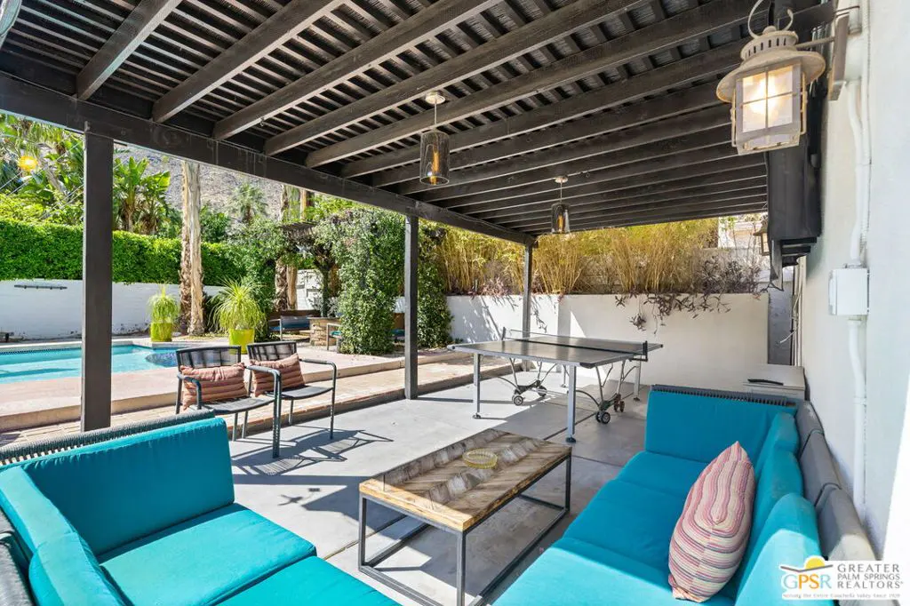Outdoor patio with blue cushioned seating, a wooden coffee table, and a ping pong table under a dark wood pergola. A pool and greenery are visible in the background.