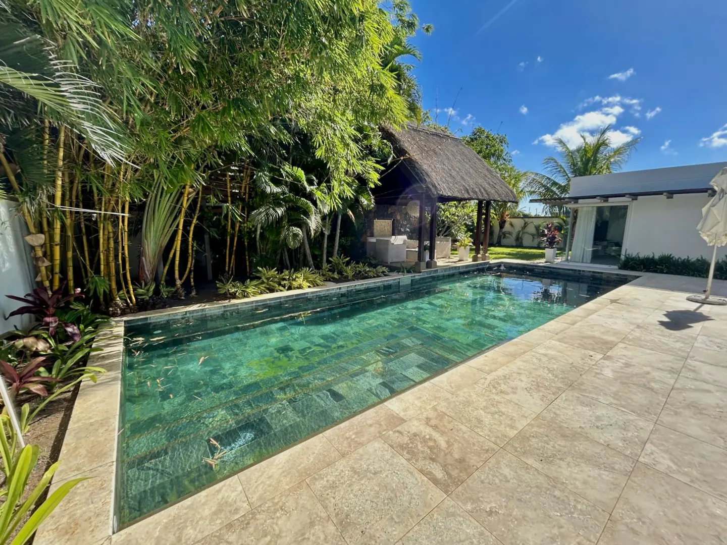 A rectangular pool with turquoise water, surrounded by beige tiles, lush greenery, and a thatched-roof gazebo.
