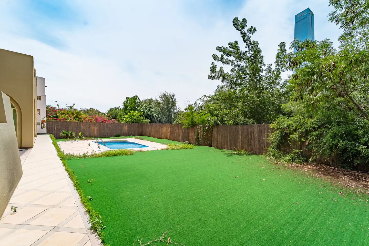 Backyard view with green lawn, blue pool, and wooden fence. Trees and a tall building are visible in the background.