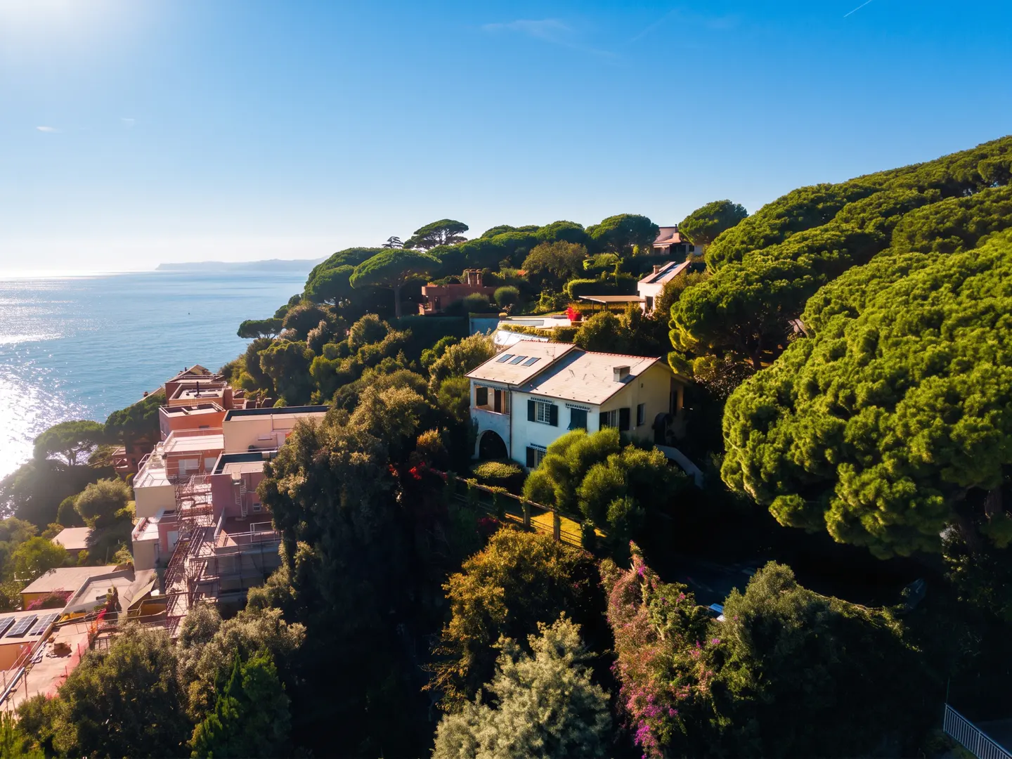 Aerial view of a white house with a red tile roof on a green, tree-covered hillside overlooking the blue ocean under a clear blue sky.