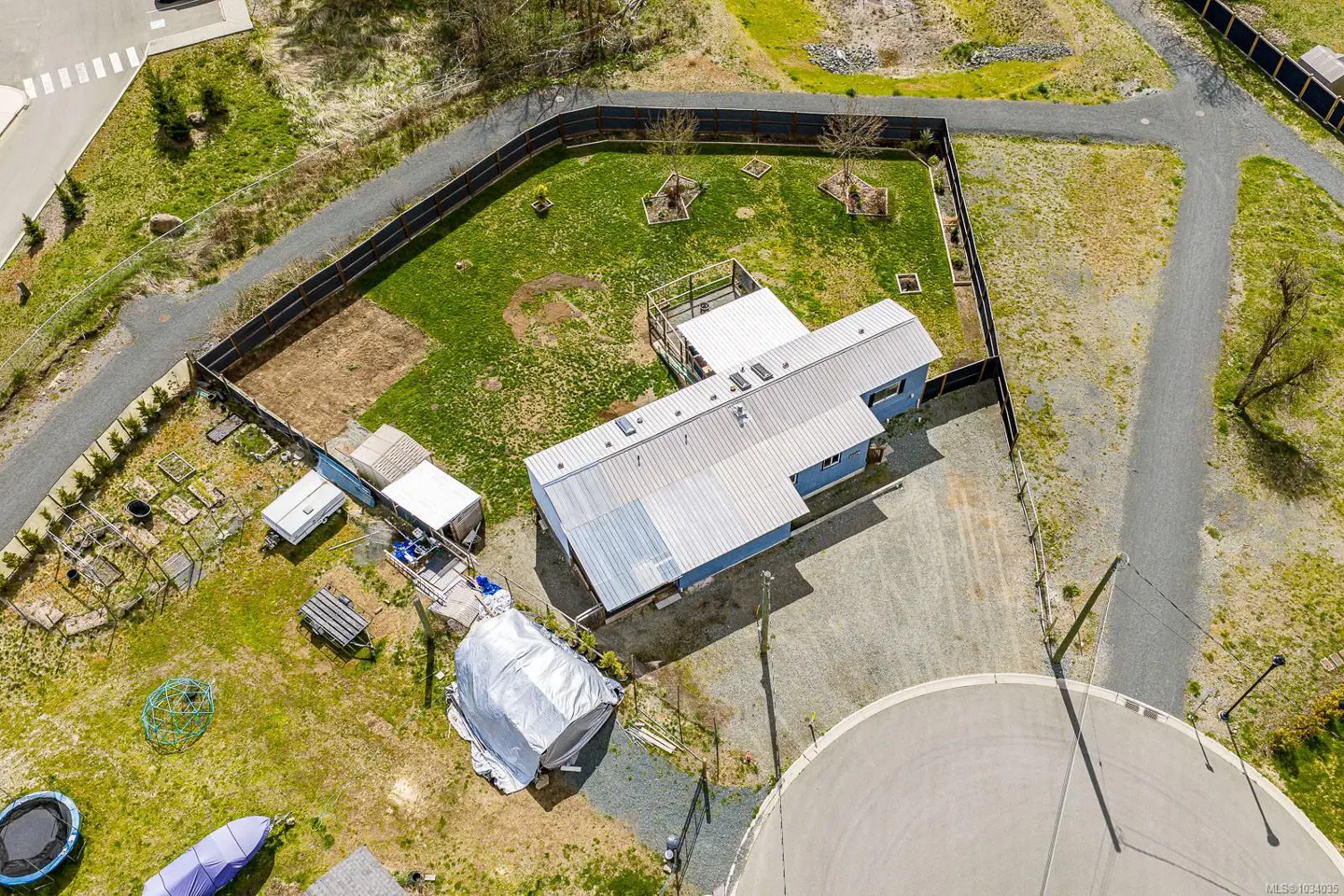 Aerial view of a blue house with a metal roof, surrounded by a fenced yard with grass, trees, and a garden. A gravel driveway leads to a cul-de-sac.