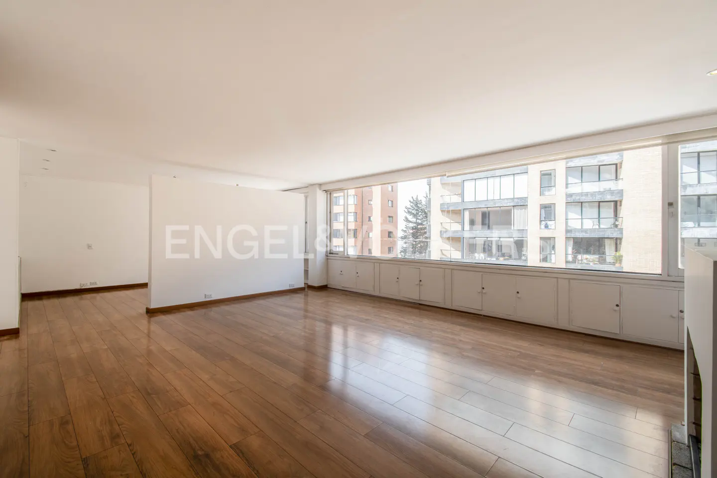 Empty living room with hardwood floors, white walls, and a large window overlooking a building.