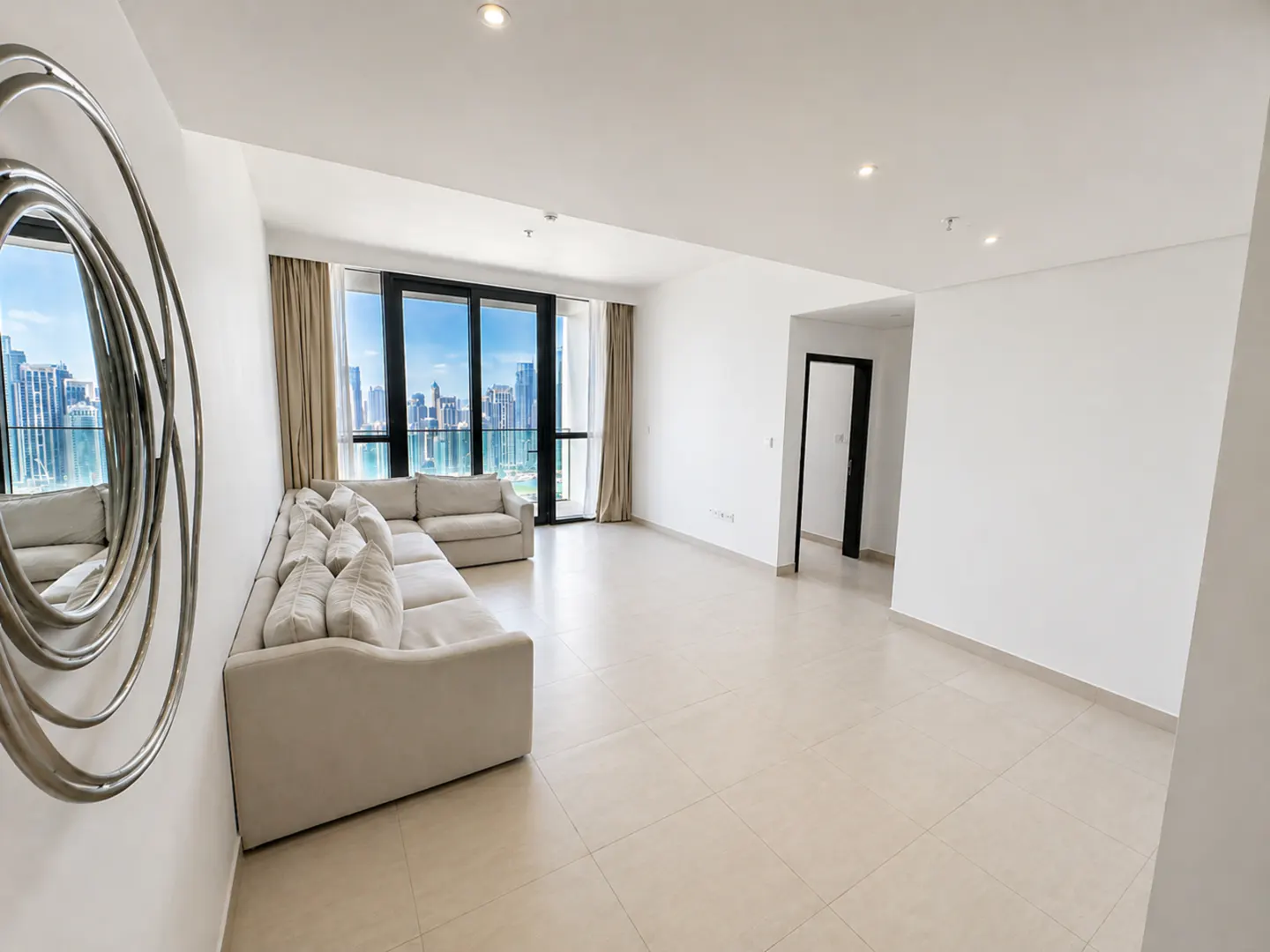 Bright living room with beige tile floor, white walls, and a large sectional sofa. A modern mirror hangs on the wall. City skyline visible through the windows.