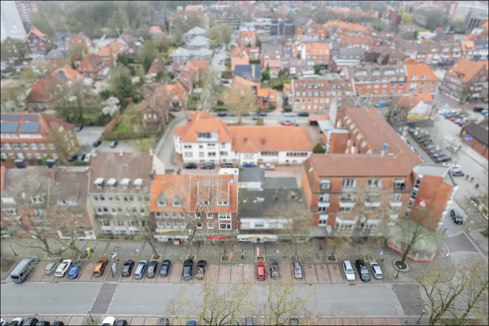 Aerial view of a European town with red-tiled roofs, buildings, trees, and cars parked along the street.